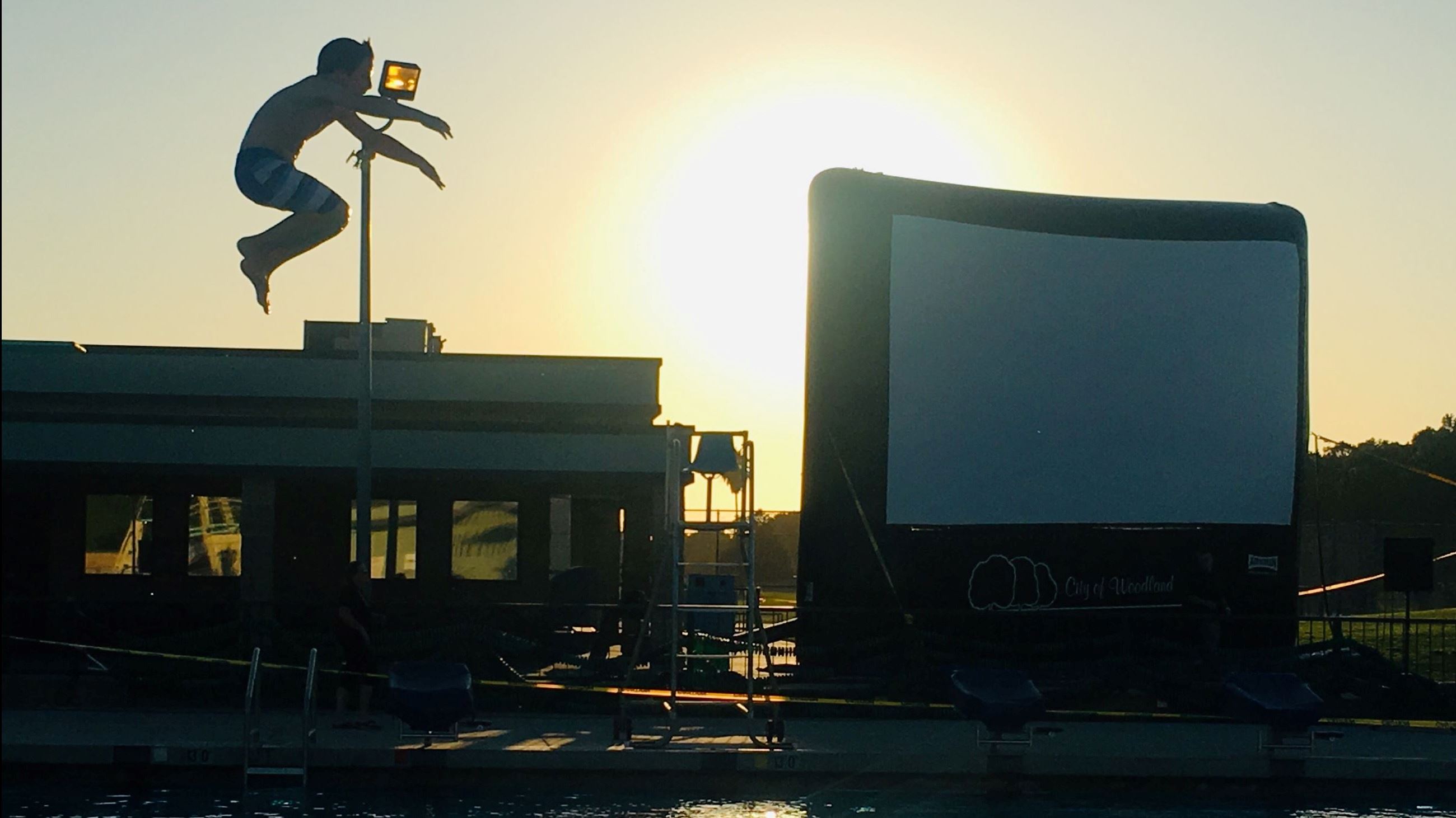 Person jumping off diving board into pool movie screen in background